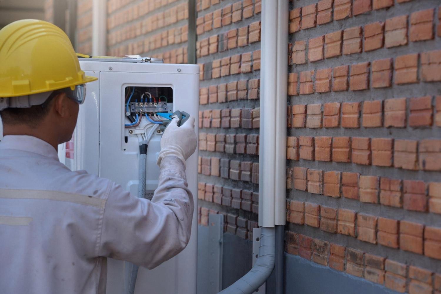 Engineer in a hard hat servicing the wiring of an outdoor air conditioner unit against a brick wall